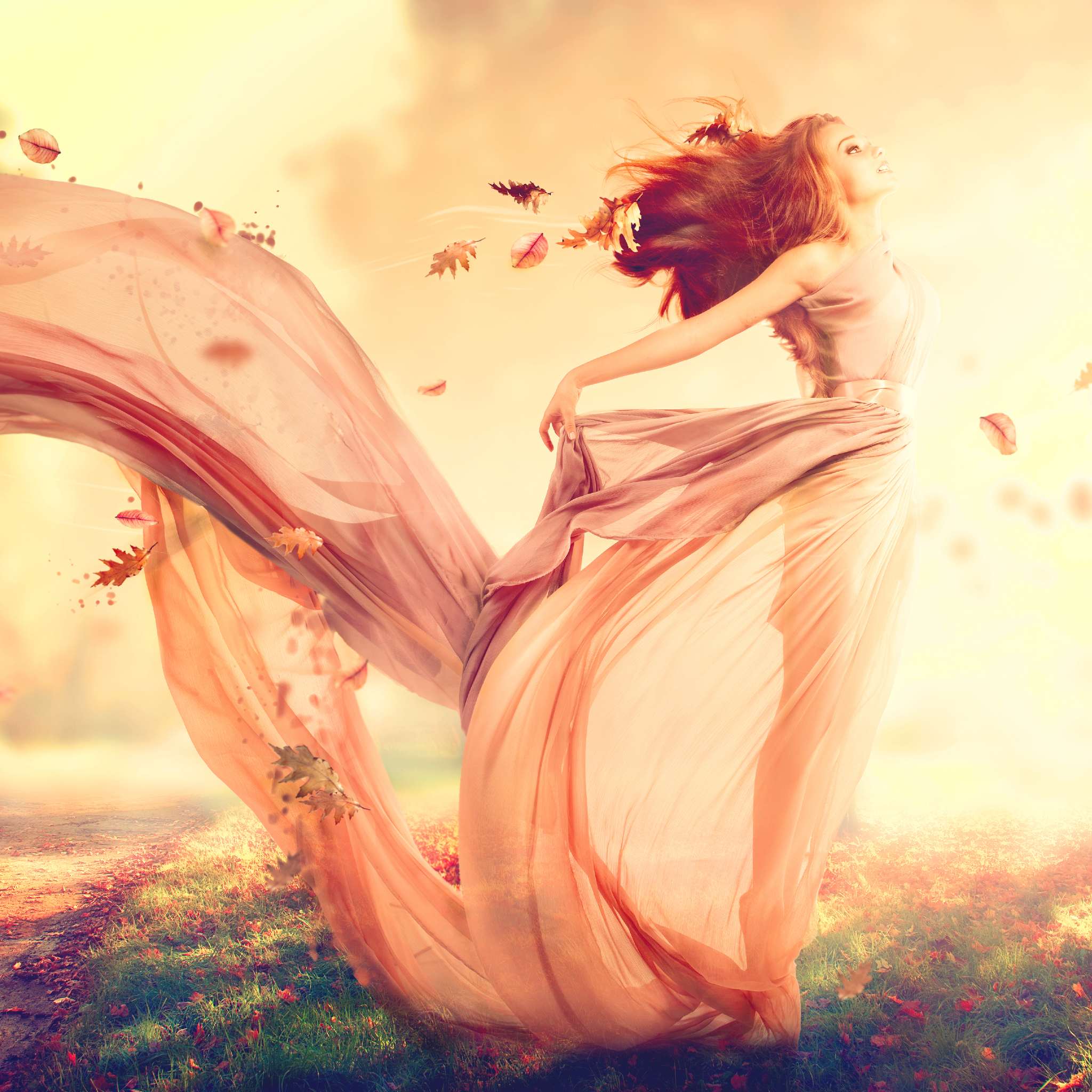 A woman with long red hair, wearing a flowing, sheer pink dress that catches the wind dramatically. She is standing in a field of green and gold grass with autumn leaves blowing around her, representing dynamic weather. The sky is a warm, bright golden yellow and orange.