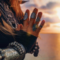 A close-up of a person's hands, heavily adorned with silver rings, bracelets, and cuffs against a warm, sunset-lit sky.