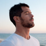 Man with closed eyes and an ear to the side standing peacefully on a beach, facing the setting sun and ocean.