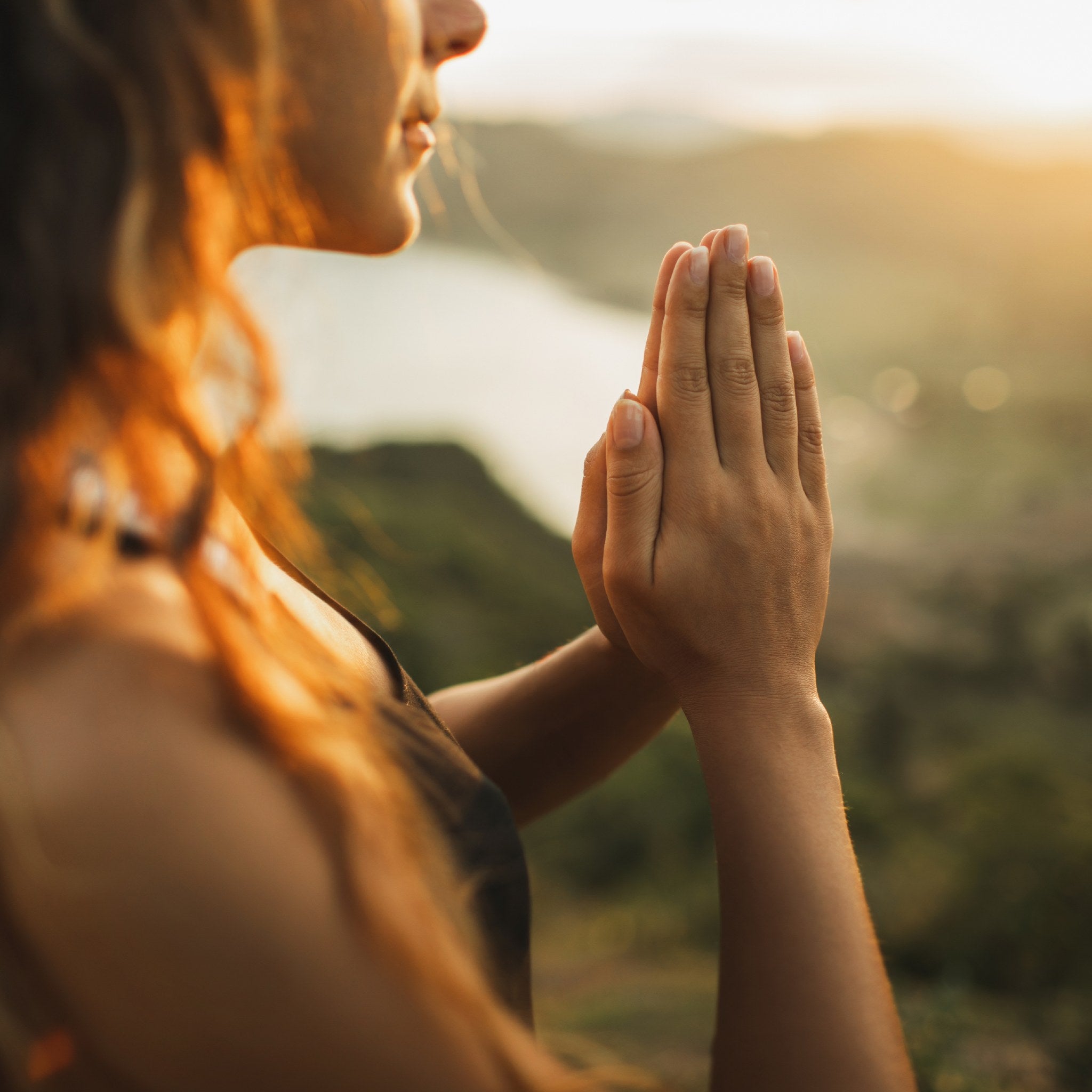 A close-up side profile of a woman meditating with her hands pressed together in a prayer pose, outdoors on a grassy hillside overlooking a valley and lake at sunset, with warm golden light illuminating her hands and hair.