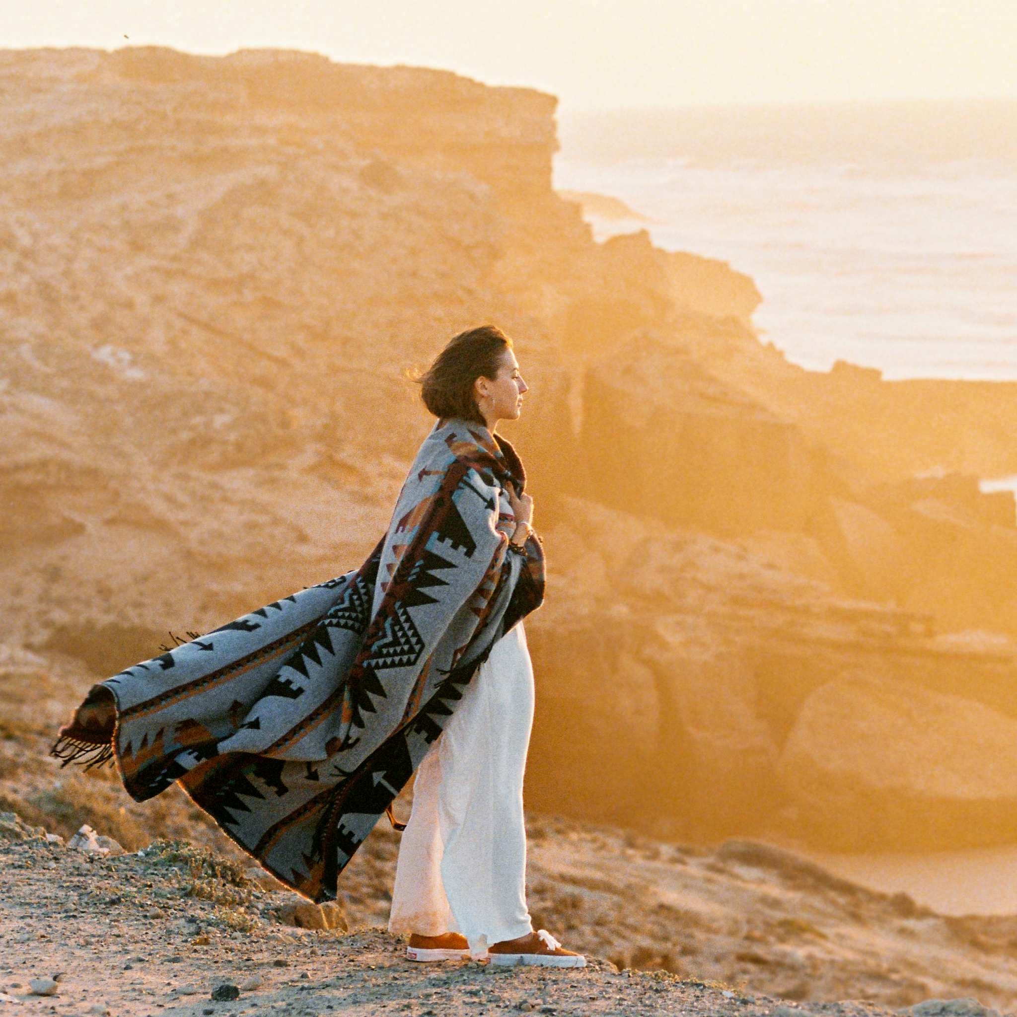A beautiful, sunlit photograph of a person wrapped in a patterned, native-style blanket, standing on a sandy cliff edge overlooking the ocean. The strong, golden light of the setting or rising sun illuminates the rugged coastal cliffs, signifying a deep connection to nature and the energy of the earth healer path alignment dedication.