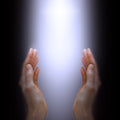 A pair of cupped, open hands reaching up from the bottom of a dark frame, cradling a bright, glowing, white-to-light-purple column of light. The image suggests the receiving or channelling of spiritual energy, illustrating an energy healer performing a treatment.