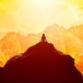 Silhouette of a person meditating on a mountain peak against a glowing orange and yellow sunset landscape.