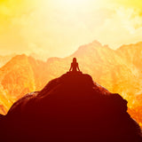 Silhouette of a person meditating on a mountain peak against a glowing orange and yellow sunset landscape.