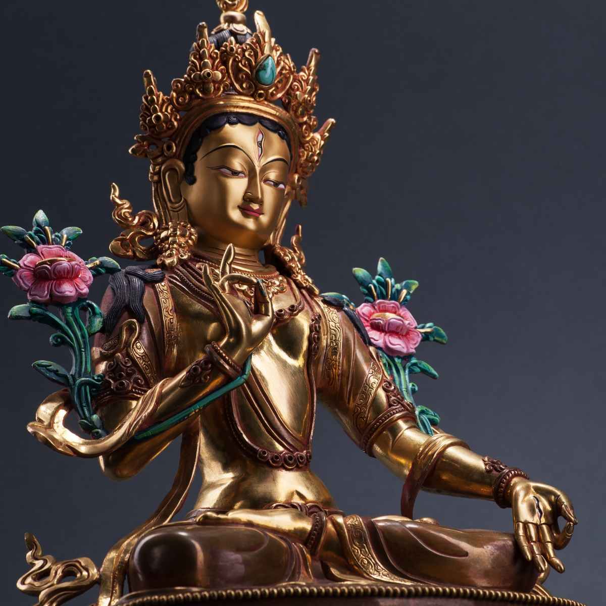 Golden statue of Green Tara wearing an ornate crown and holding pink lotus stems against a dark background.
