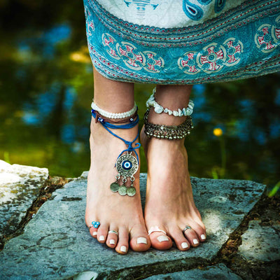 A close-up photograph of a person's bare feet, standing on a grey stone slab near the edge of a pond. The ankles are adorned with multiple beaded and metal anklets, including a striking round charm with a blue evil eye symbol, representing the connection of the grounding chakras to the earth.