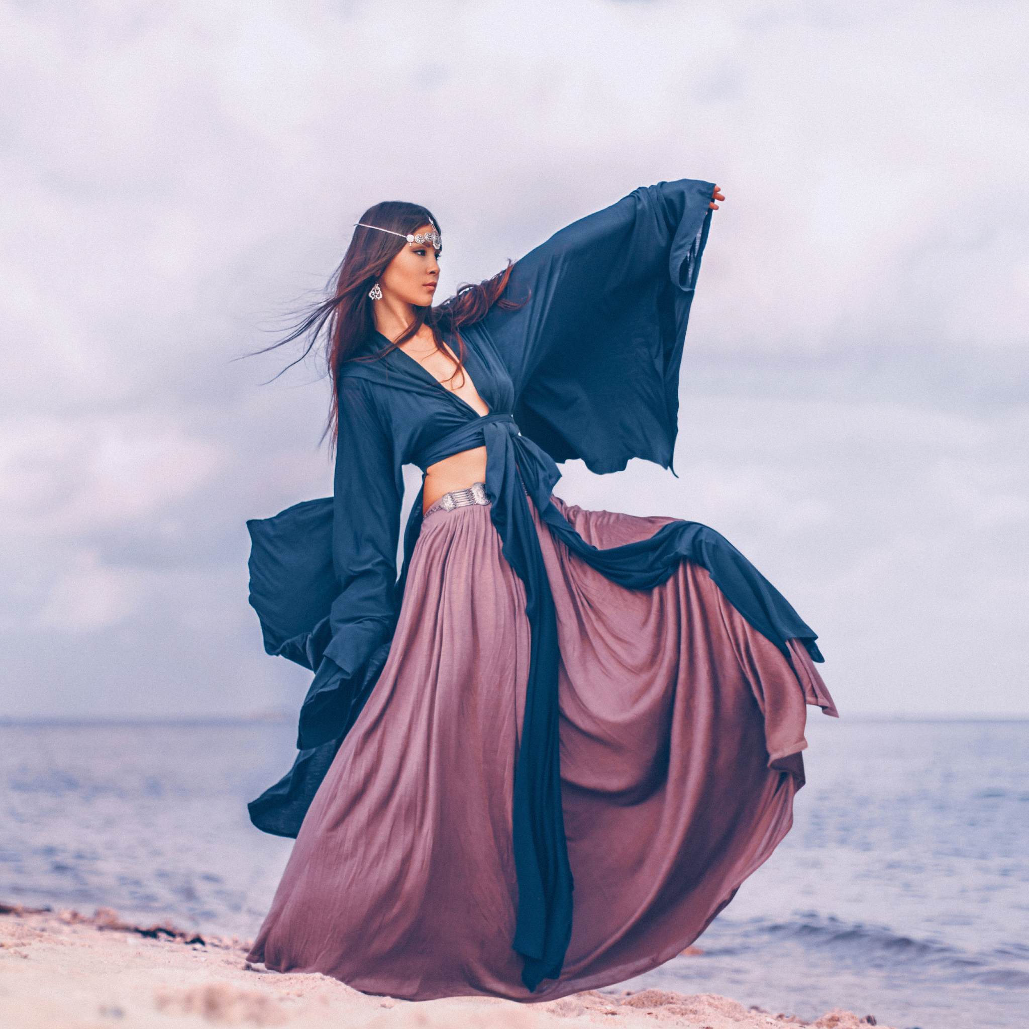 A person standing on a beach with long, flowing clothing in shades of purple and blue, with the ocean and sky in the background.