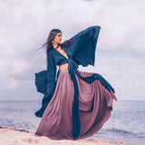 Woman in flowing clothing in shades of purple and blue on a beach with a cloudy sky.