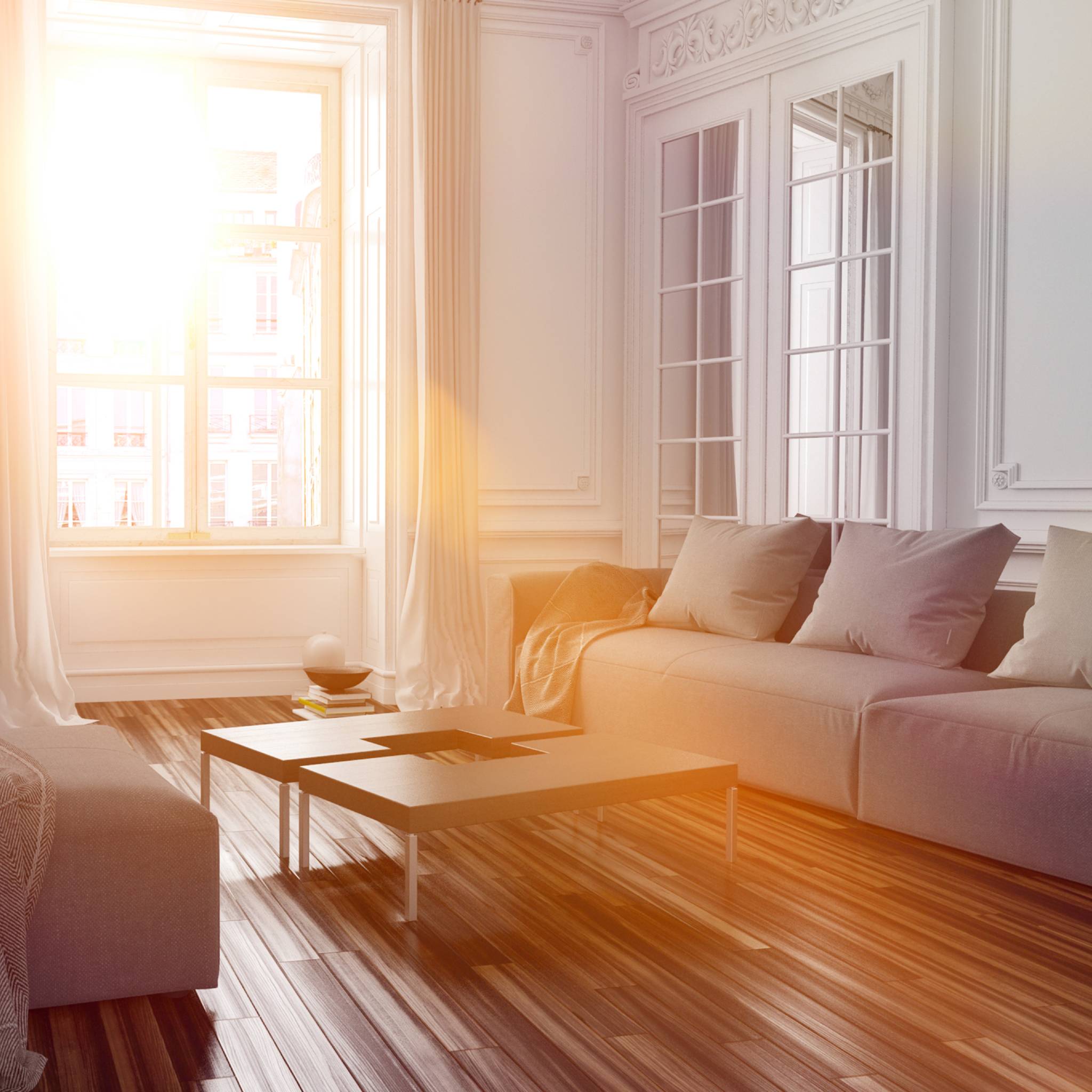 A bright, sunlit modern living room with tall white walls, elegant molding, and a plush grey couch, featuring a large window with white curtains and a view of city buildings, with a warm sun flare filling the image.