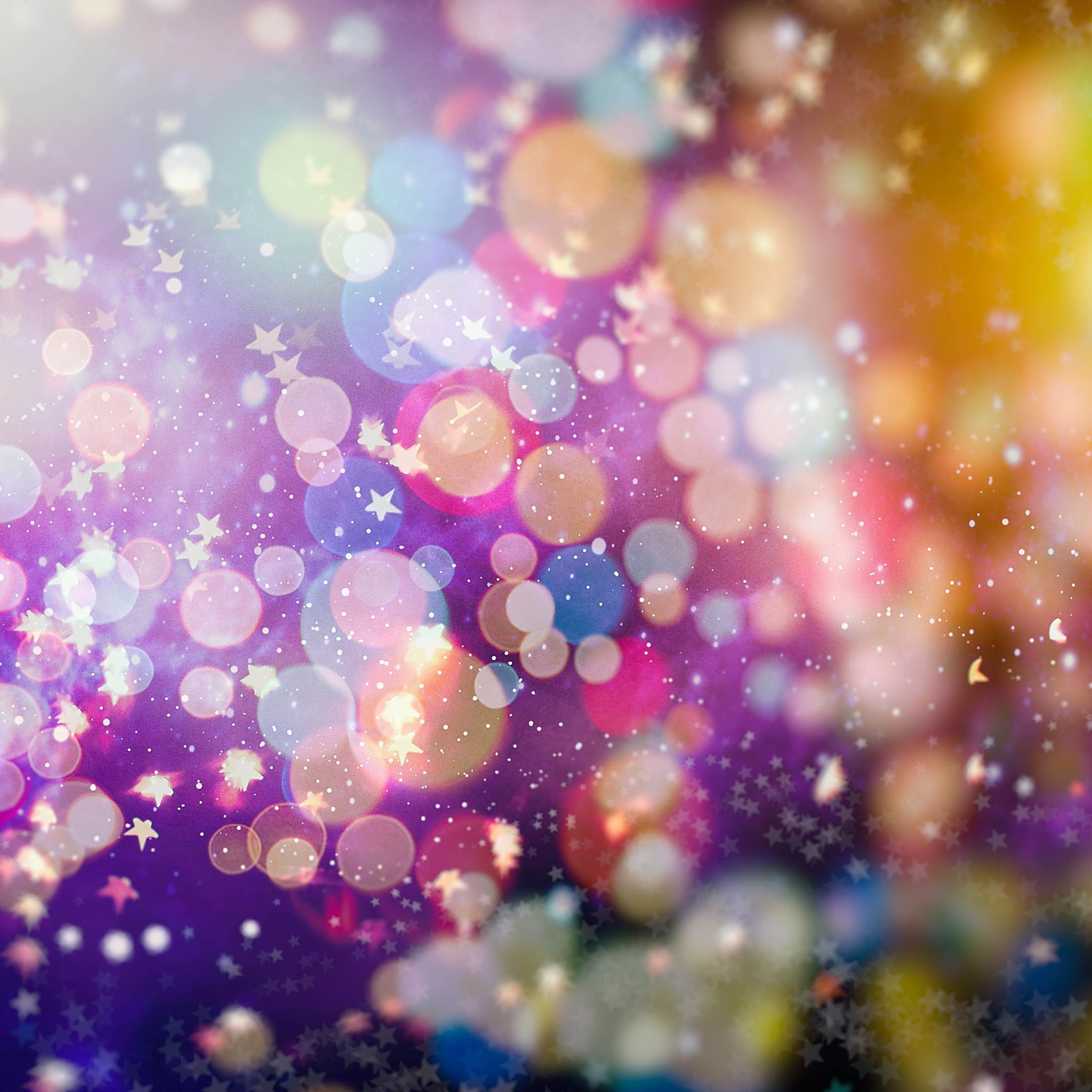 An abstract, brightly coloured bokeh background featuring large, soft circles of pink, purple, yellow, and blue light, with numerous small white stars or sparkles floating throughout the foreground.
