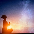 A dark silhouette of a person sitting in a cross-legged position with hands in a prayer pose, looking upward. The figure is brightly backlit by an intense orange and yellow glowing light at the base, contrasting with a deep blue, starry cosmic sky above.