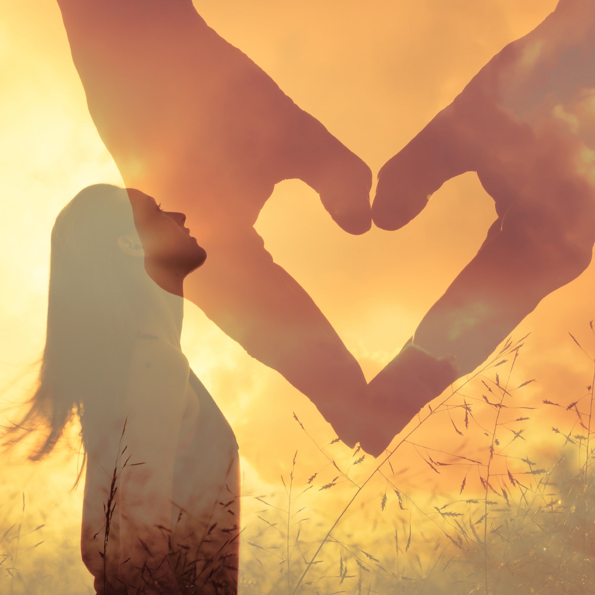 Silhouette of a woman looking up into the sky with a double-exposed heart shape made out of two hands against a sunset sky.