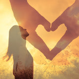 Silhouette of a woman looking up into the sky with a double-exposed heart shape made out of two hands against a sunset sky.
