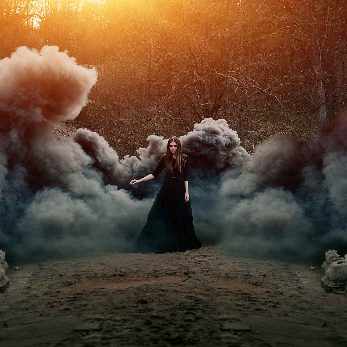 Woman in a long black dress standing amidst dramatic plumes of dark smoke in a sunlit forest.