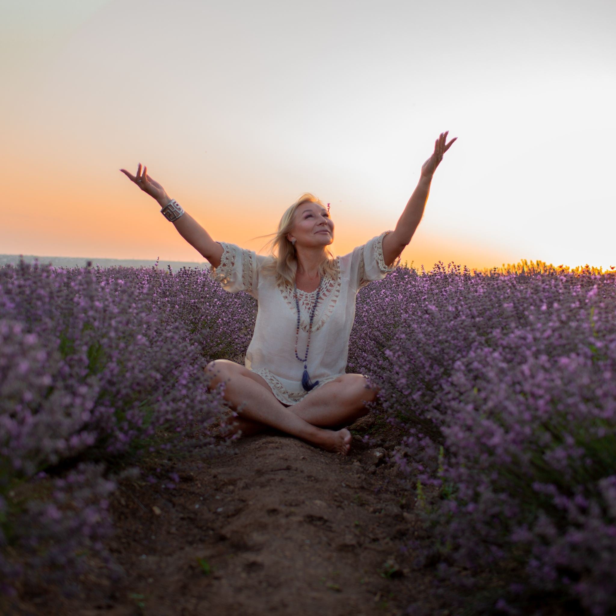 A photograph of a blonde woman sitting barefoot and cross-legged in a vast, purple lavender field at sunset, with her arms raised to the sky in a posture of joyful acceptance and prayer. She is smiling and looking upwards, representing the alignment and intention of the manifestation path alignment dedication.