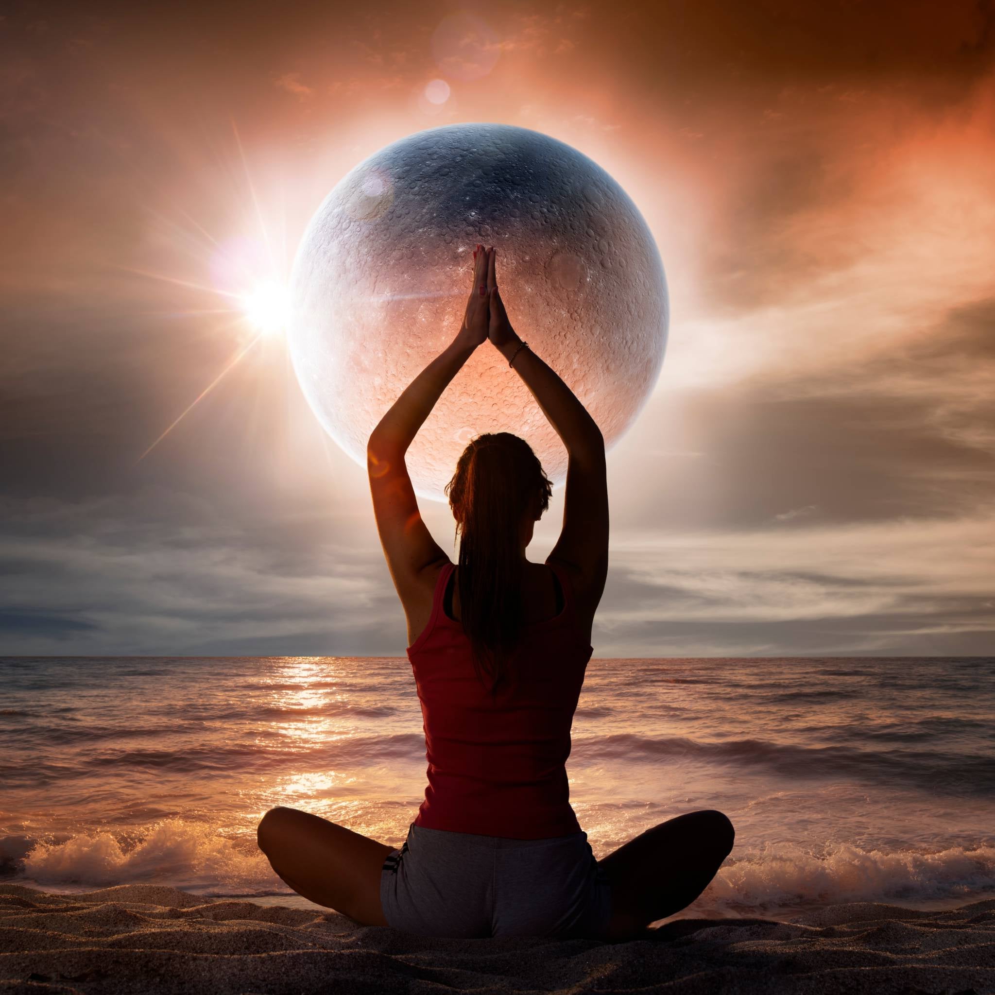 A photograph of a person in a red vest and shorts sitting cross-legged on a beach at sunset, with their back to the camera and hands clasped above their head in a prayer pose. A large, dramatic, textured moon and a bright sun flare dominate the sky over the shimmering ocean.