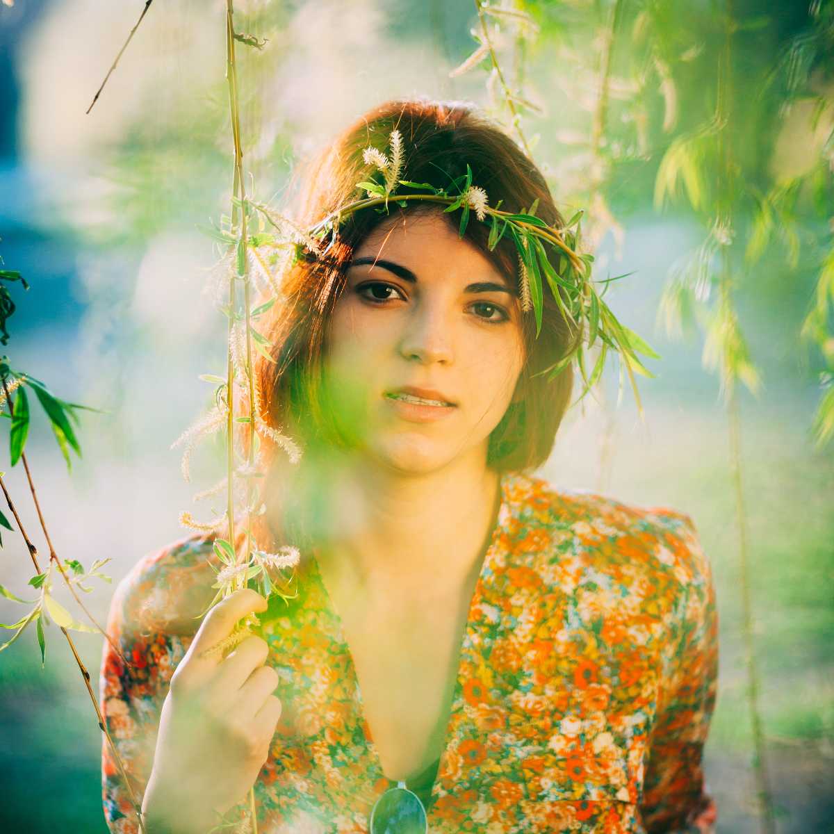 Young woman with a leafy crown wearing a floral dress amidst weeping willow branches in sunlit nature.