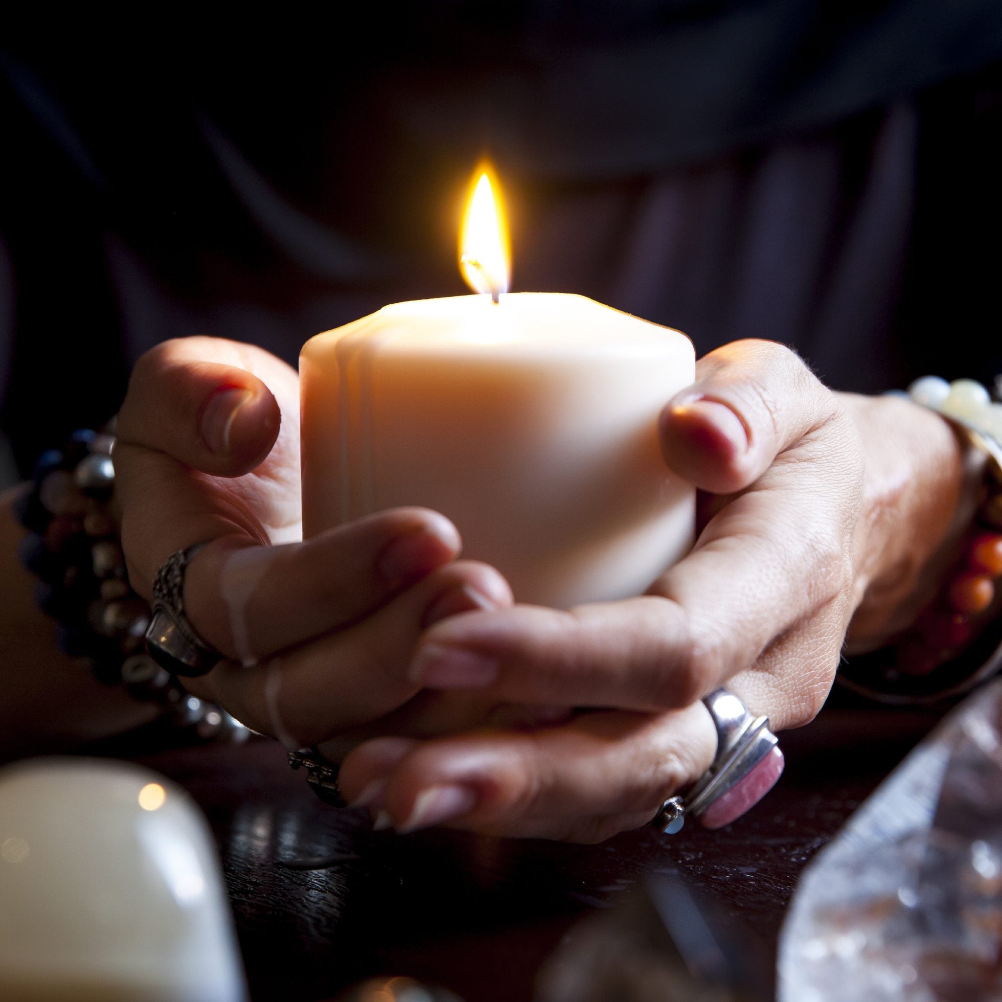 Close-up of hands wearing jewellery and bracelets, cupping a thick, lit white pillar candle in a dark setting.