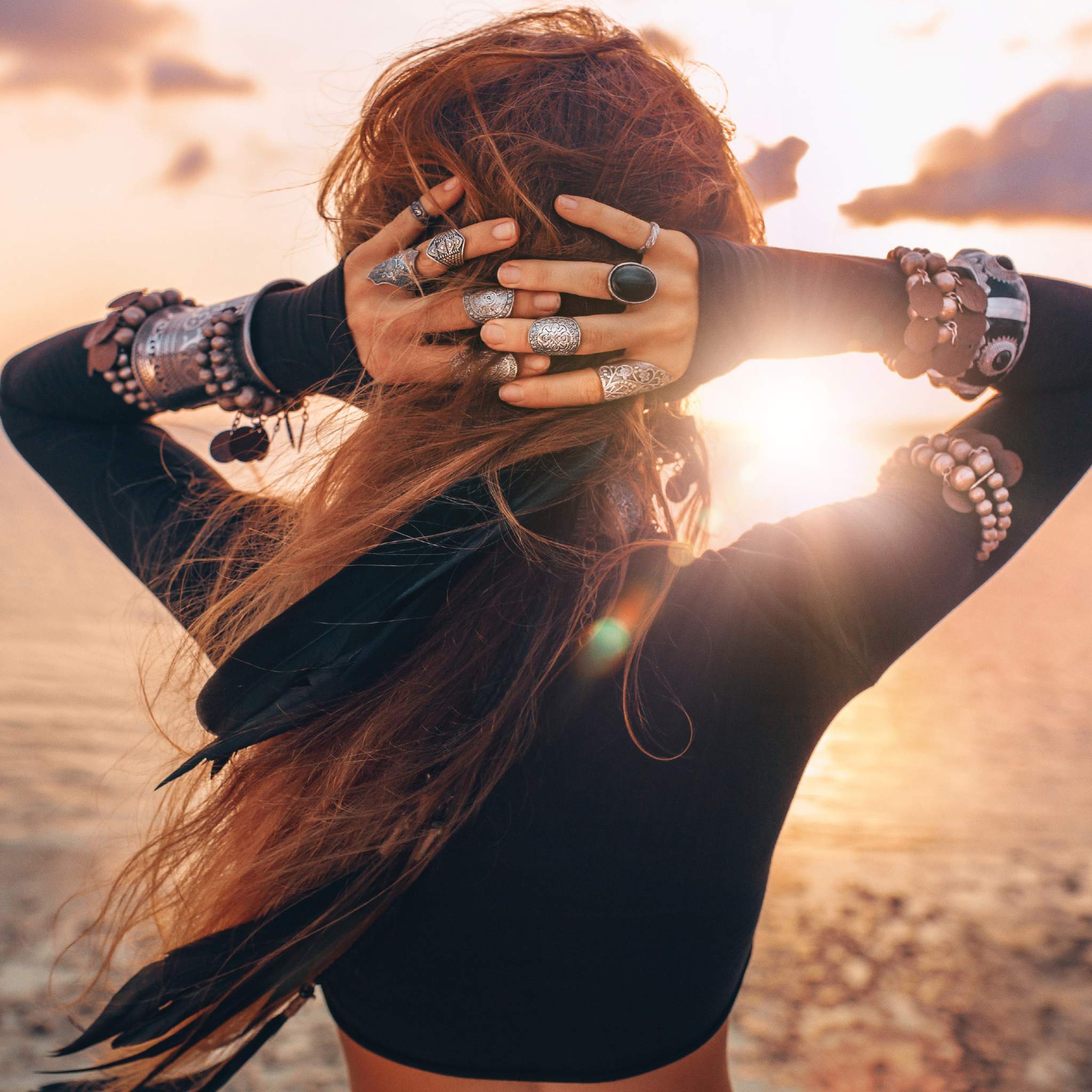 A woman with hands behind her head, adorned with rings and bracelets, facing a sunset over water, representing psychic Lemurian spiritual awareness.