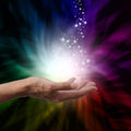A close-up photograph of an outstretched hand, palm up, with a brilliant, cascading stream of white light and sparks falling into it. The background is a dramatic, swirling vortex of rainbow colours in a smoky, ethereal effect, representing the healing energy channelled by a reiki practitioner.