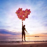 A silhouette of a person jumping on a beach at sunset, holding onto a cluster of pink and red balloons shaped like a large heart. The sun sets over the ocean, casting an orange and pink glow on the waves and the sand, illustrating themes of the inner child and love.