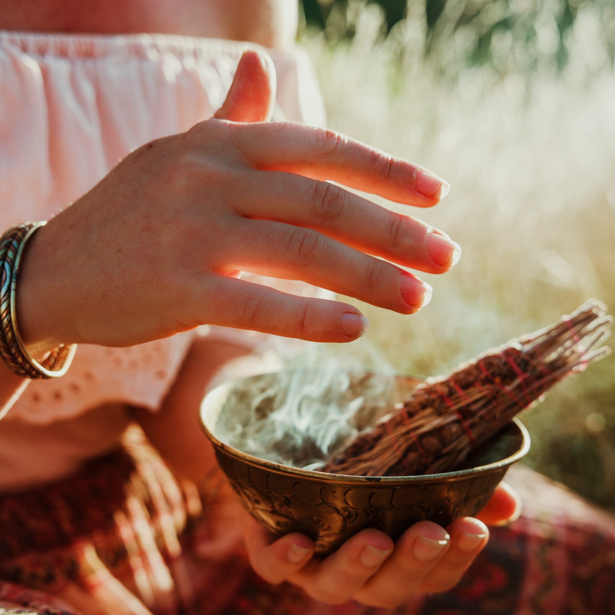 A close-up, sunlit photograph of a person's hands holding a small, ornate bronze bowl from which a thin stream of smoke rises, signifying burning sage or a similar cleansing herb bundle wrapped in twine. One hand is held slightly above the bowl, interacting with the smoke, symbolising the act of purifying a space for a space clearer path alignment dedication.