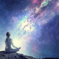 Silhouette of a figure sitting in a yoga or meditation pose on a rocky outcrop, looking up at a vast, bright, purple and blue cosmic galaxy.