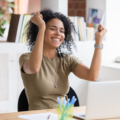 A photograph of a young woman with curly dark hair sitting at a desk in a bright office, smiling broadly at the camera with her hands raised in fists of triumph. She is celebrating a victory at her laptop, symbolising successful business.
