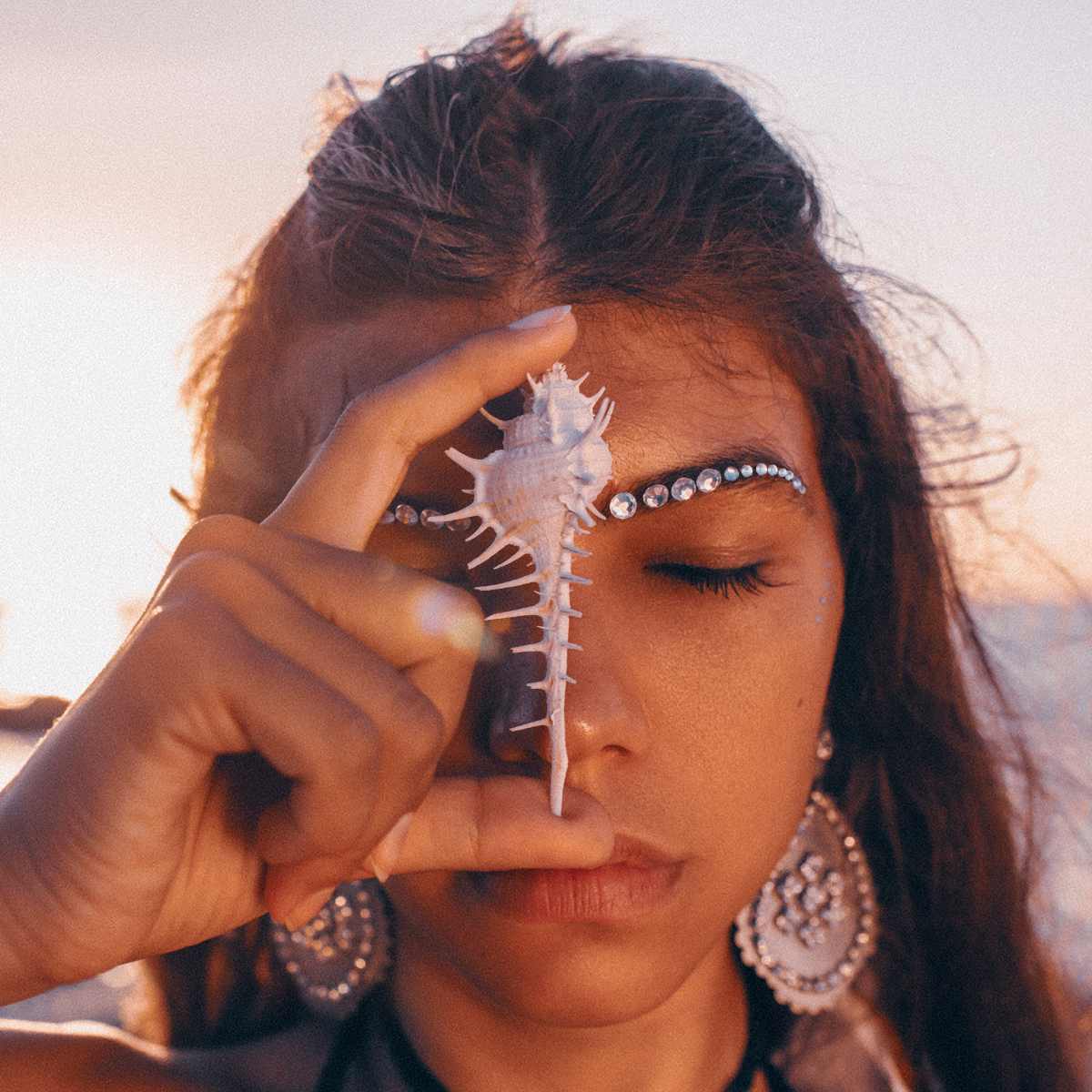 Woman holding a shell to her third eye with a blurred beach background.