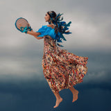A woman in a colourful dress with a blue feathered headdress rising up against a cloudy sky.