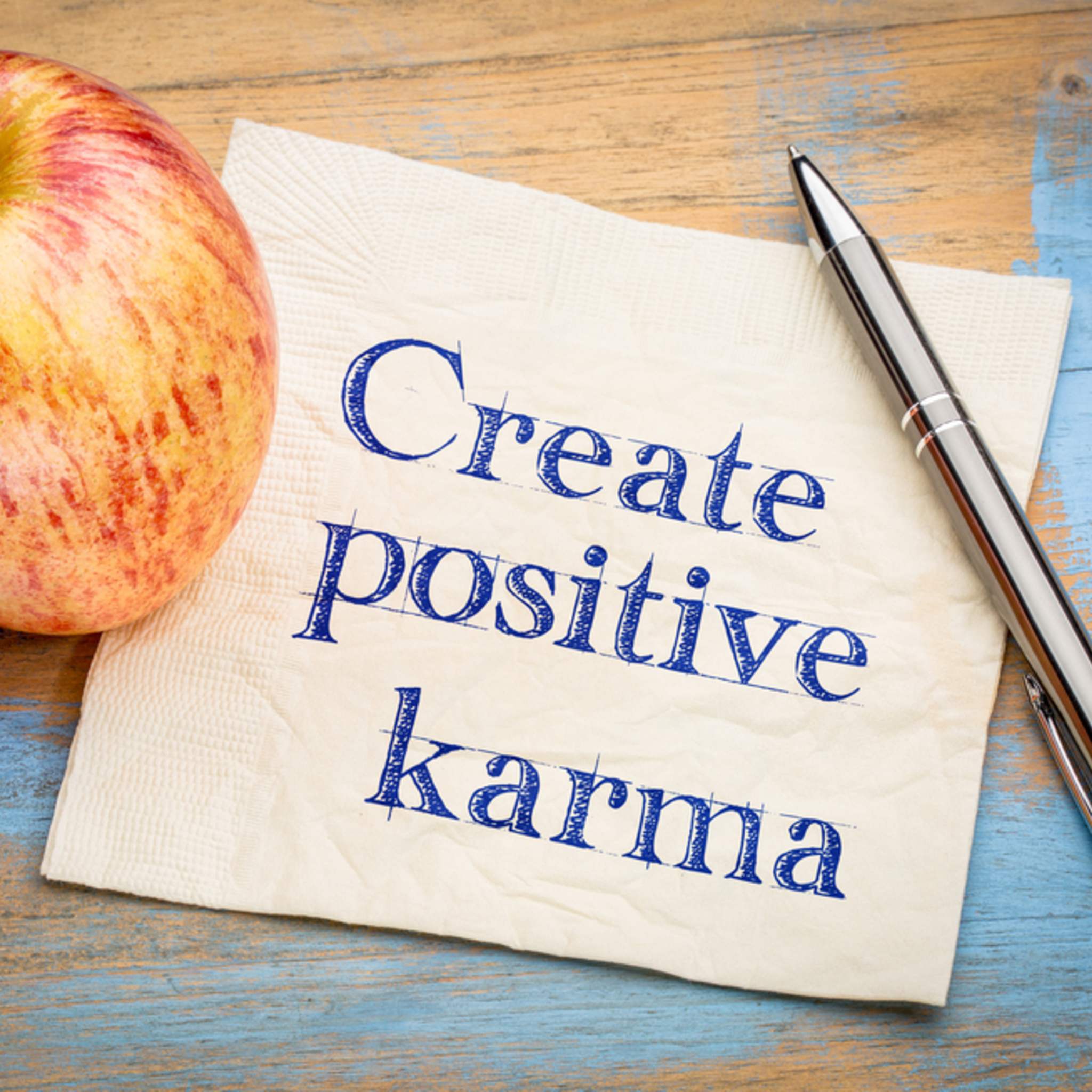 A white napkin with 'Create positive karma' handwritten in blue, next to a red apple and a silver pen, on a blue rustic wooden background. Symbolises conscious creation of positive outcomes.