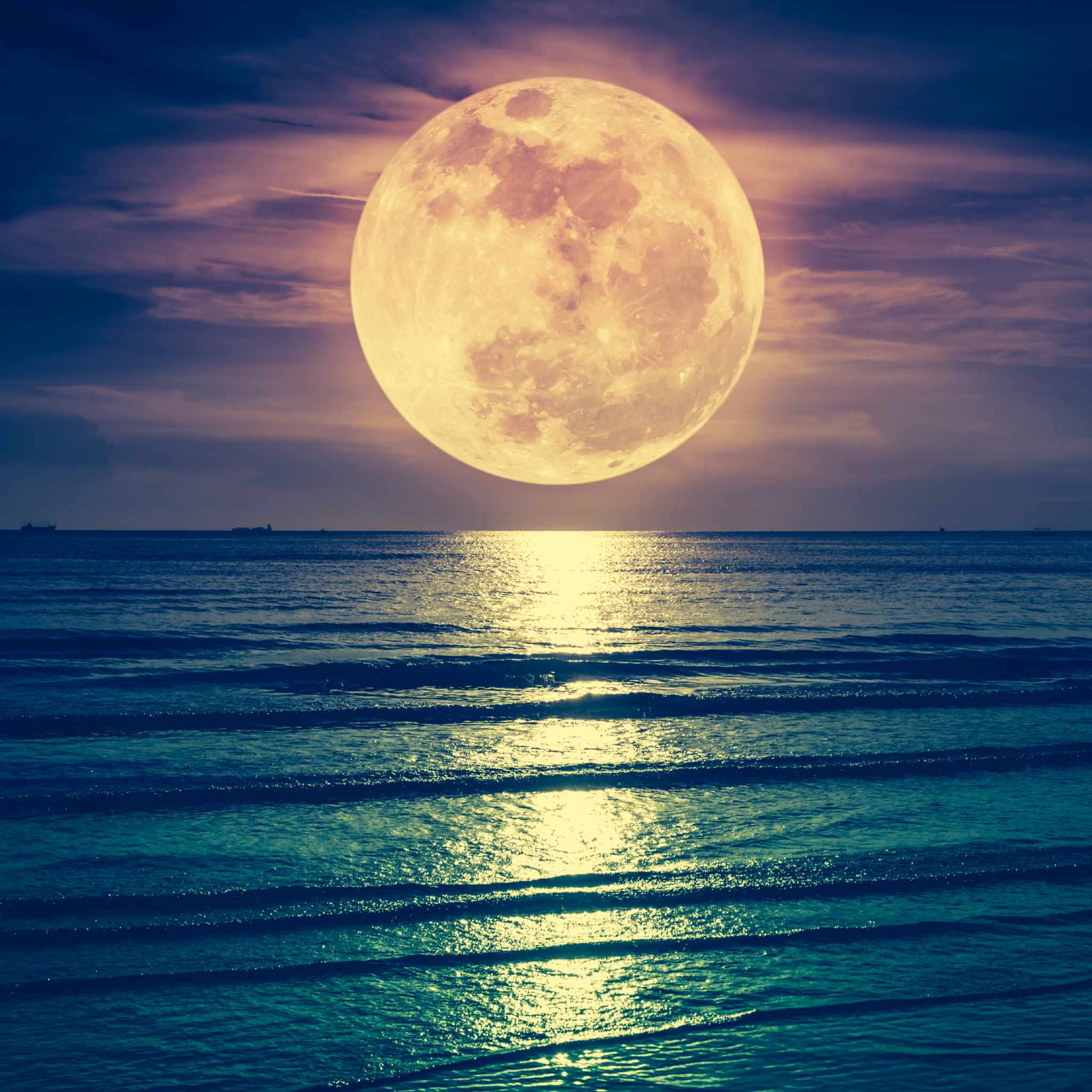 A striking nighttime photograph of a massive, glowing orange full moon suspended low over a teal-coloured ocean. The moon's light reflects brightly across the water in a vertical path, with faint ships visible on the distant horizon, representing lunar illumination healing.