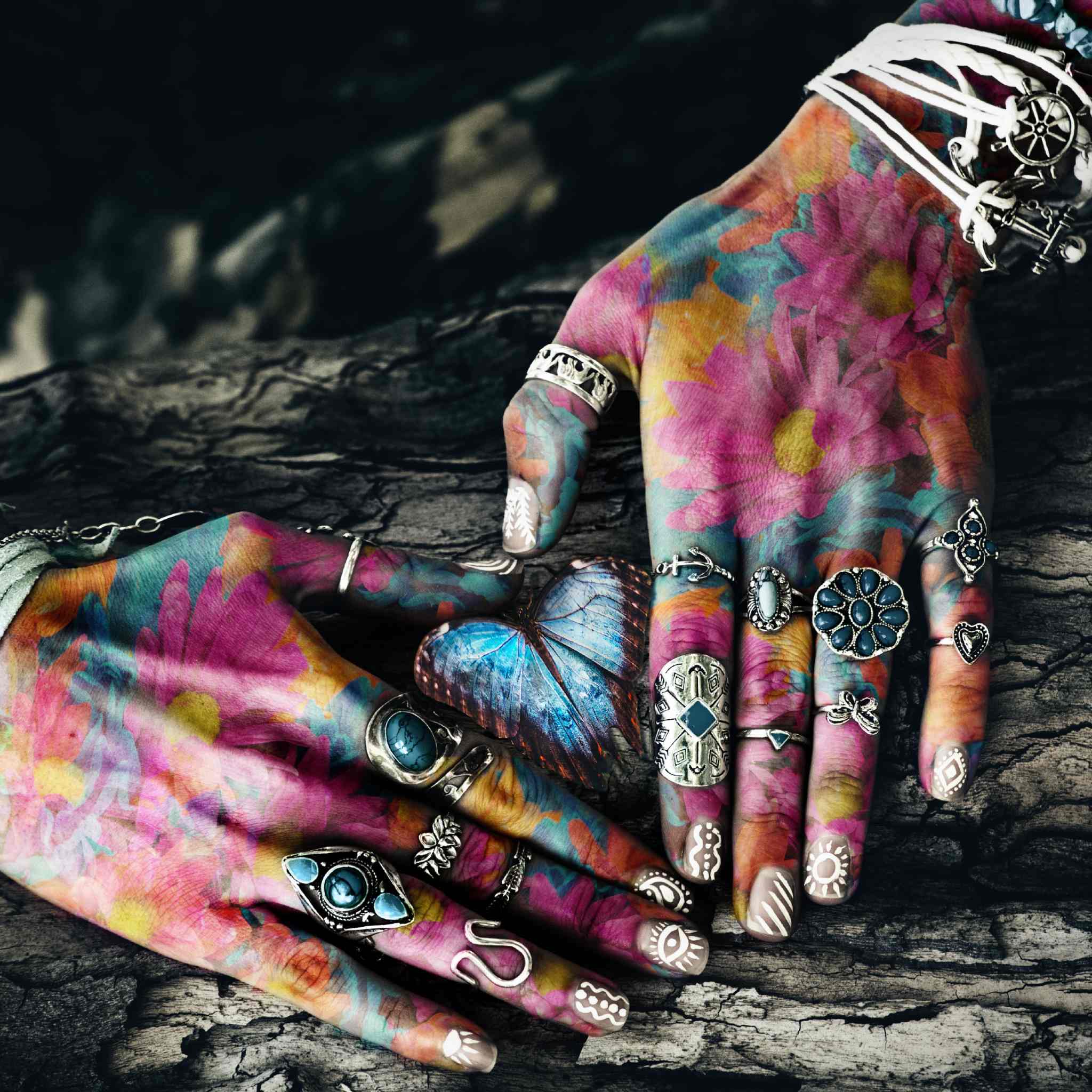 A close-up of two hands resting on dark, rough bark. The skin is painted in a vibrant, multi-coloured floral design (pink, yellow, and blue), and both hands are heavily adorned with silver and turquoise rings and bracelets. A large, iridescent blue butterfly rests between the hands, illustrating a theme of ancestral honouring.