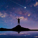 A silhouette of a person standing on a rock with their arms outstretched, looking up at a starry night sky with a bright shooting star and a reflection in the water.
