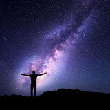 A silhouette of a person standing on a hill with their arms outstretched, gazing up at a vibrant purple and blue Milky Way galaxy in the night sky.