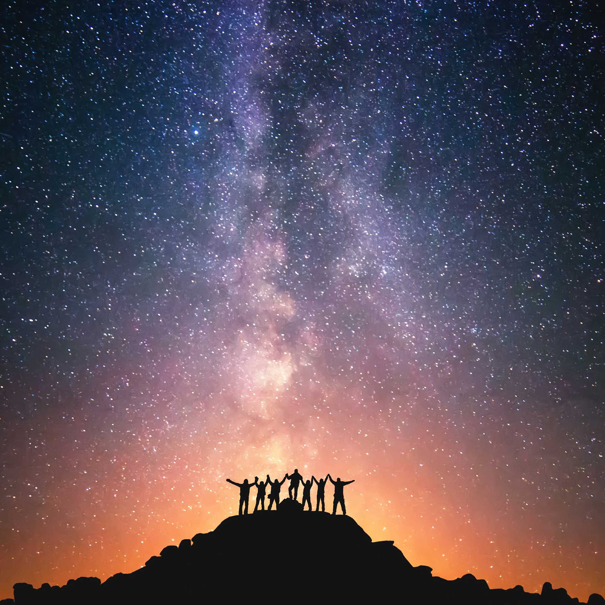 A silhouette of a group of seven people standing on a hill holding hands under a magnificent starry night sky with the Milky Way.