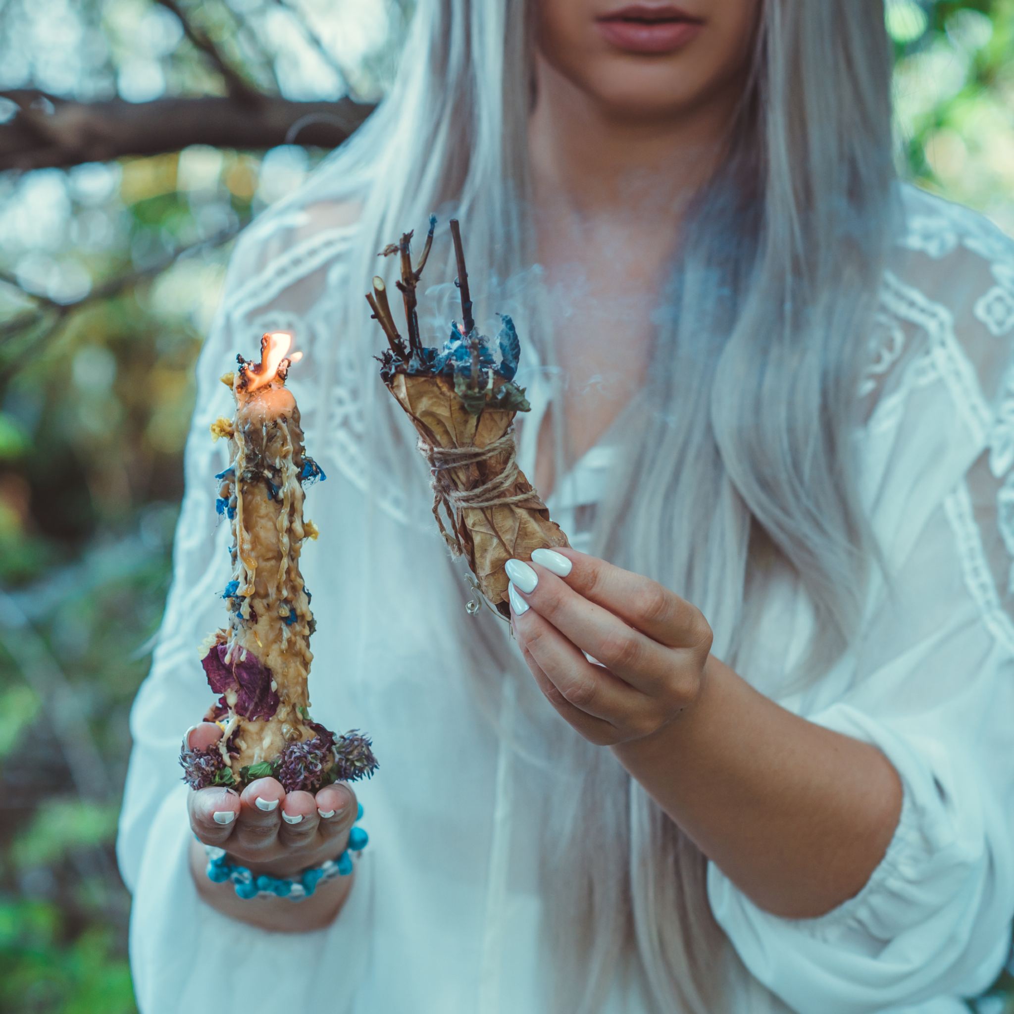 A close-up photograph of a person with long, white-blonde hair and a white lace blouse, holding a large, handmade, decorated, burning beeswax candle in their left hand and a bundle of smoking ritual herbs and sticks in their right hand. The image is set outdoors amongst green foliage, symbolising the cleansing and spiritual focus of the white magic path alignment dedication.
