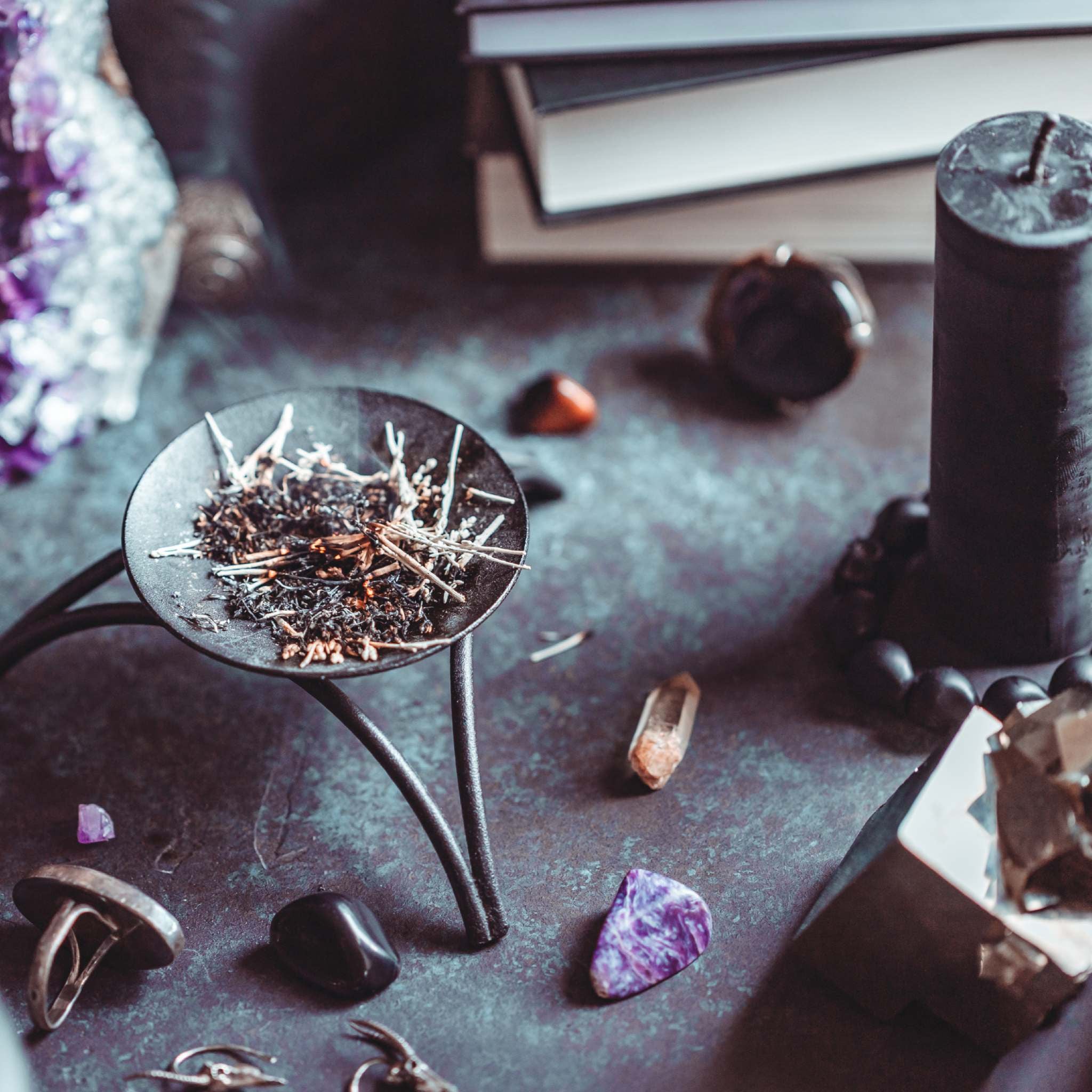 A close-up, dimly lit photo of a witch's ritual altar surface covered in various tools, including a black cast iron incense burner on a tripod stand filled with loose herbs, a tall black candle, polished gemstones, pieces of raw crystal, and stacked books. This collection of items represents the spiritual focus of the witch path alignment dedication.